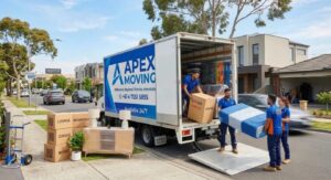 Apex Moving residential moving service in Melbourne, showing team in blue and orange uniforms packing fragile items, a wooden crate with a painting, and labeled boxes including KITCHEN-1 and LIVING-2. An open moving truck is visible through the door with the city skyline in the distance.